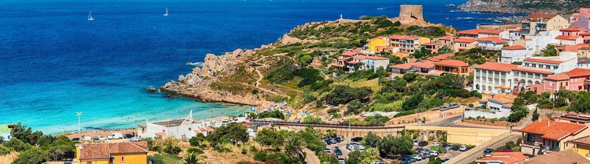 Küstenlandschaft auf Sardinien mit türkisblauen Meer rechts und bunten Häusern links, darunter Parkplätze. Ein sandiger Strand mit Badenden und bunte Sonnenschirme erstrecken sich in der Mitte.