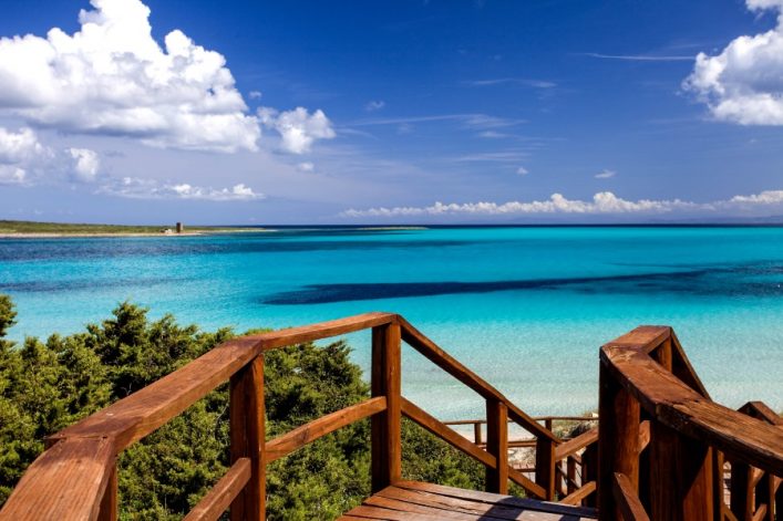 Holzgeländer einer Treppe führt zur türkisblauen Küste, Horizont mit weißen Wolken, dichter Strauch im Vordergrund links, alter Turm sichtbar auf Landzunge links im Hintergrund.