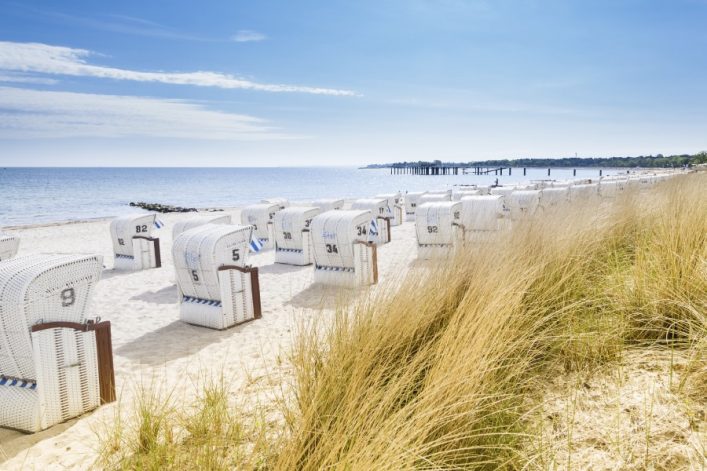 Strand mit weißen Strandkörben am Ufer, dahinter das blaue Meer. Links Sand, rechts hohe Gräser. Im Hintergrund ein langer Steg. Klarer Himmel mit wenigen Wolken.
