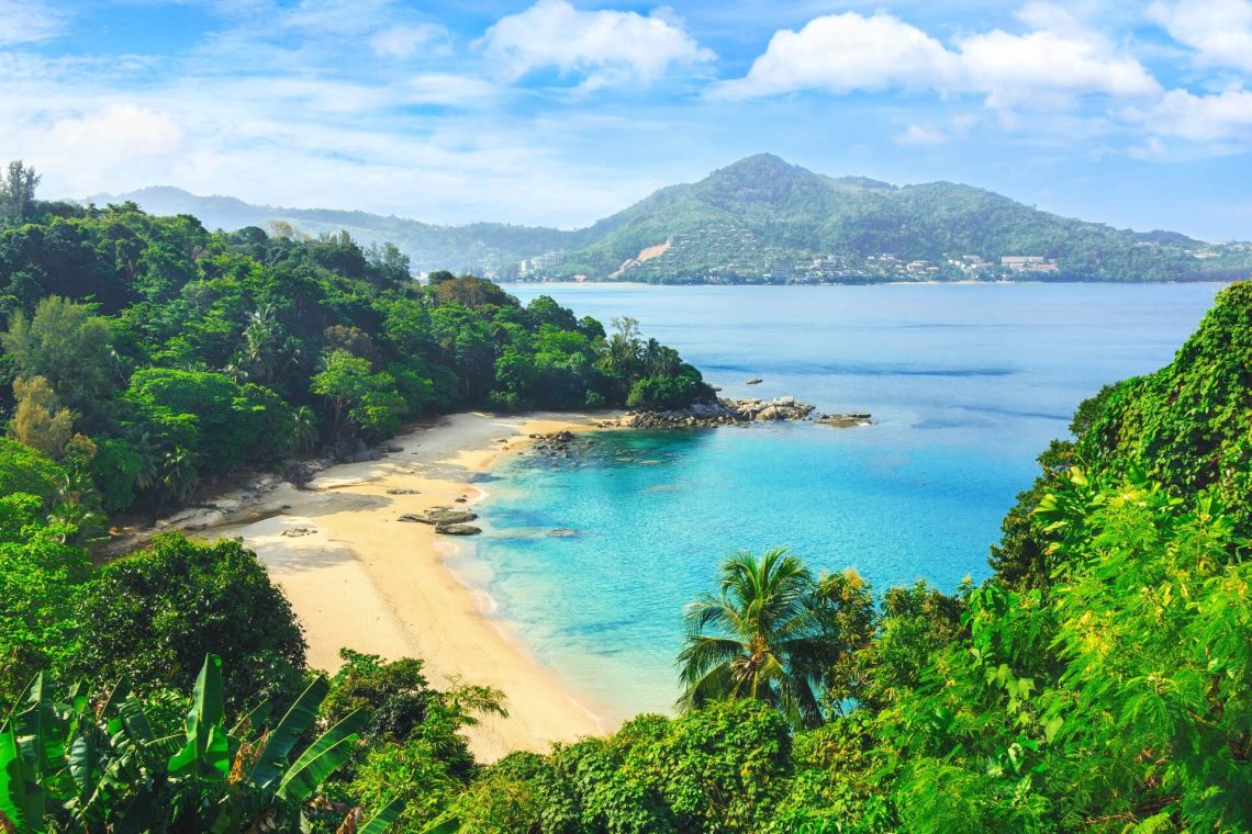 Tropische Bucht in Phuket mit goldenem Sandstrand, umgeben von üppigem Grün und klarem, türkisfarbenem Wasser – ideal für Ferien am Meer in Thailand.