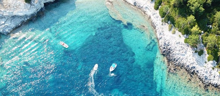 Klares, türkisfarbenes Wasser umspült die Felsen und das grüne Ufer der griechischen Insel Paxos. Mehrere Boote gleiten auf dem Wasser. Im Hintergrund dichter Wald an der felsigen Küste, die halbkreisförmig das Meer umrahmt.