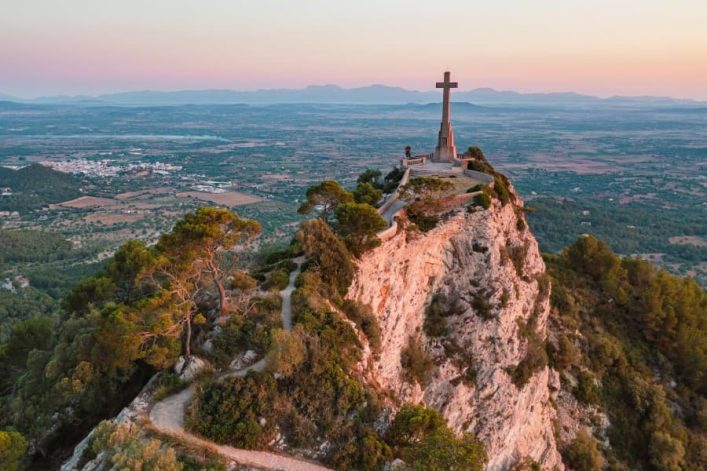 Sonnenaufgang beleuchtet Sant Salvador auf Mallorca: Kreuz auf Klippe, Serpentinenweg führt hinauf, weite Aussicht über die Ebene im Hintergrund.