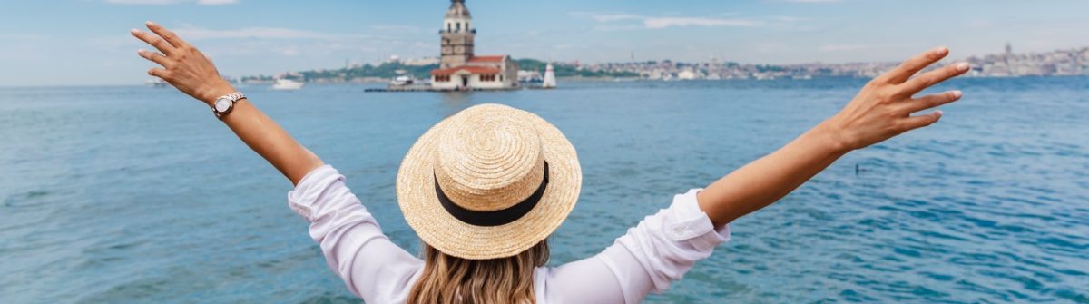 Frau mit Strohhut breitet vor dem Bosporus die Arme aus, Rücken zur Kamera, blickt auf den historischen Mädchenturm, eine Perle Istanbuls.