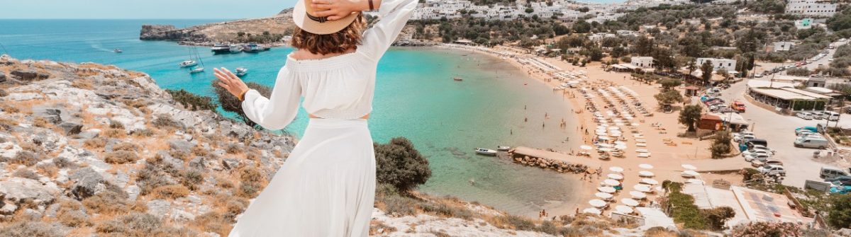 Frau in Weiss blickt auf Lindos mit seiner Akropolis auf Rhodos, umgeben von azurblauem Meer und sandigem Strand.