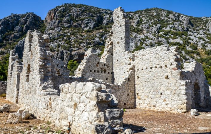 Ruinen der antiken Stadt Olympos bei Antalya mit weiß-grauen Steinmauern, die teils verfallen sind. Im Hintergrund ragen bewaldete, felsige Hügel in den strahlend blauen Himmel empor.