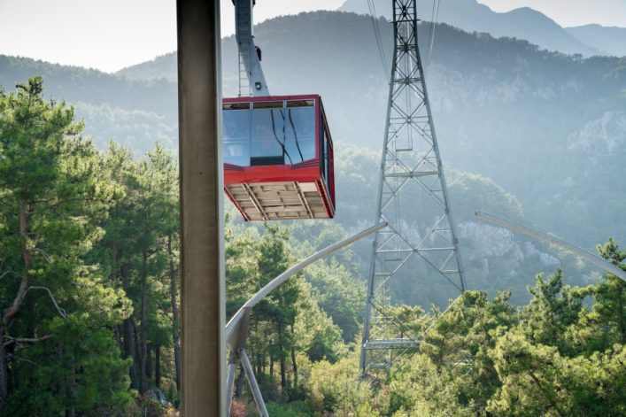 Eine rote Seilbahngondel schwebt über einem Waldgebiet, eingerahmt von großen Nadelbäumen. Rechts davon steht ein hoher Stahlturm, der die Seile trägt. Im Hintergrund erheben sich die Berge Antalyas.