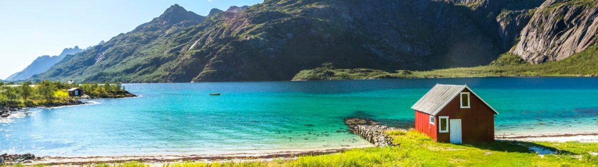 Eine idyllische Küstenszene in Norwegen mit einer roten Hütte rechts im Vordergrund, klarem türkisblauem Wasser des Trollfjords, umgeben von grünen Wiesen und eindrucksvollen Bergen im Hintergrund.