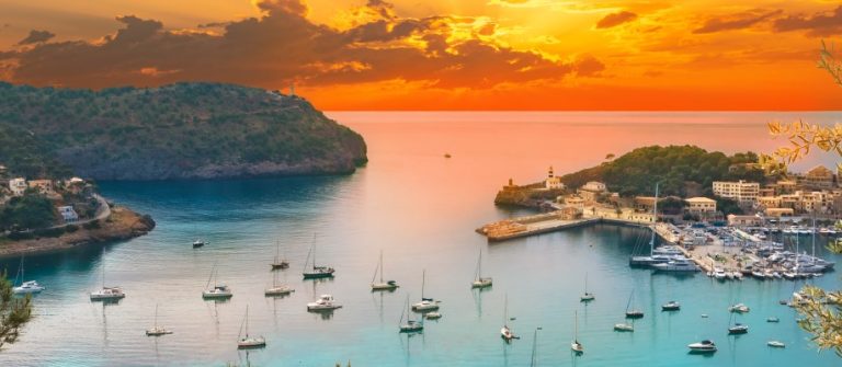 Sonnenuntergang über Port Soller, Mallorca; Bucht mit mehreren Segelbooten, umgeben von Hügeln und üppigem Grün, orangefarbener Himmel spiegelt sich im ruhigen Wasser, Gebäude am Ufer links und rechts.
