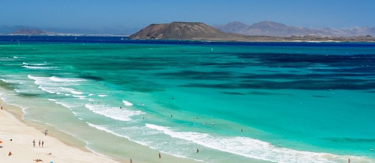 Weisser Sandstrand mit zahlreichen Menschen links, türkisblaues Meer in der Mitte, sanfte Wellen, im Hintergrund eine karge Insel unter klarem blauem Himmel.