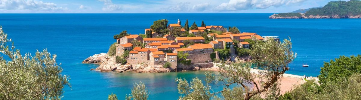 Sveti Stefan, eine malerische Insel vor Montenegro. Traditionelle Häuser mit orangefarbenen Dächern, von tiefblauem Meer umgeben. Im Vordergrund Olivenbäume, im Hintergrund klarer Himmel und Hügel in der Ferne.