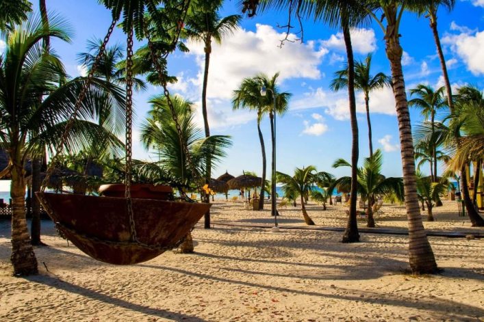 Palmen am weißen Sandstrand, blauem Himmel und reetgedeckten Sonnenschirmen im Hintergrund. Vordergrund zeigt ein großes Metallgefäß, an Ketten aufgehängt. Im Hintergrund ist das strahlend blaue Meer.