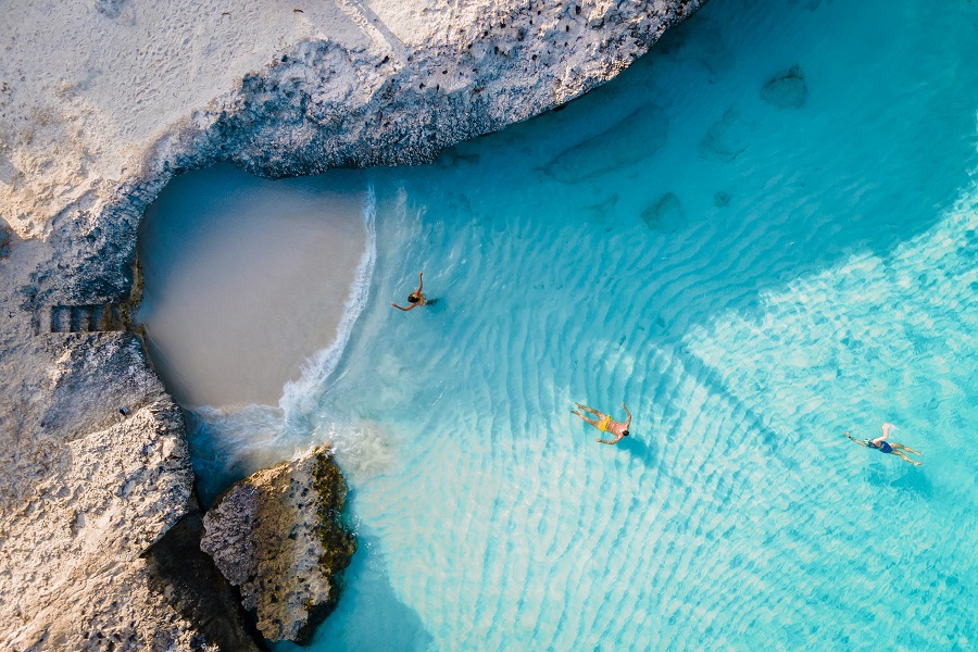 Drei Personen schwimmen im türkisblauen Meer von Tres Trapi auf Aruba. Oberhalb ist eine kleine Sandbucht von Felsen umgeben. Links führen Stufen ins Wasser, daneben ist ein großer Felsblock.