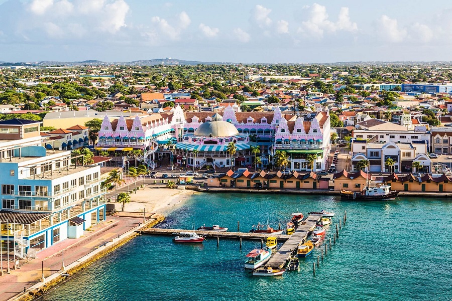 Bunte Gebäude mit rosa Fassaden und weißen Dächern im Zentrum von Oranjestad, Aruba, umgeben von kleineren Häusern und üppigem Grün. Vor dem Ufer liegen kleine Boote am Pier im türkisblauen Wasser.