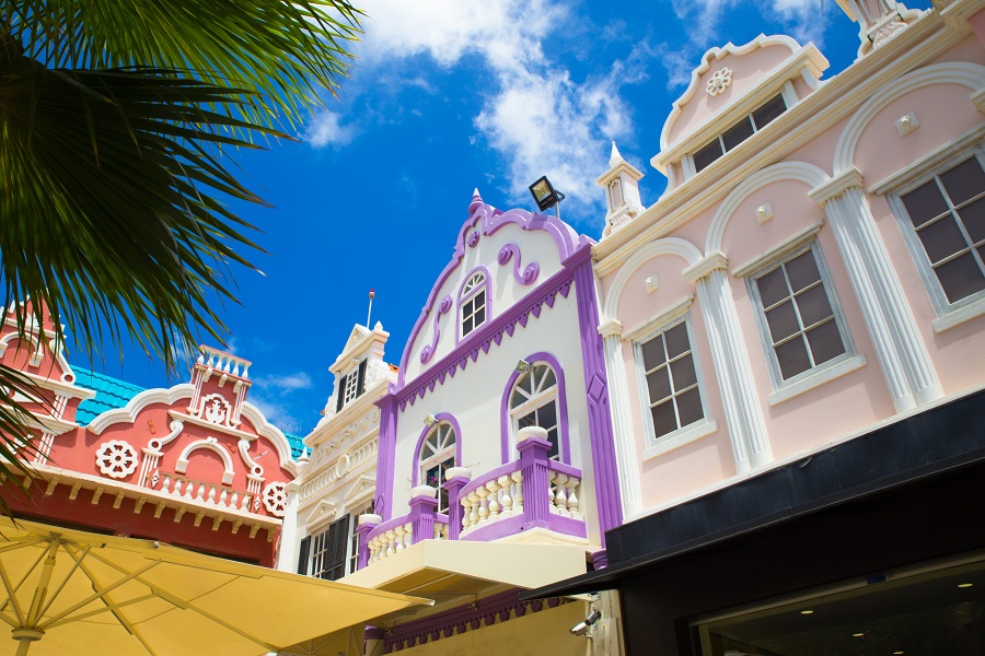 Bunte, pastellfarbene Gebäude mit aufwendigen Fassaden und holländischer Architektur in Oranjestad, Aruba. Links ragt eine große grüne Palme ins Bild, im Vordergrund ist ein gelber Sonnenschirm zu sehen und darüber der blaue Himmel mit wenigen Wolken.