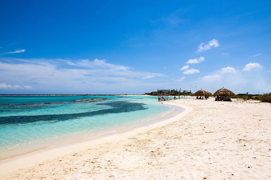 Feiner, weißer Sandstrand, türkisfarbenes Meer mit dunklen Flecken, Strohschirme rechts, einige Menschen darunter. Klare, blaue Himmel mit wenigen Wolken im Hintergrund. Tropisches Flair.