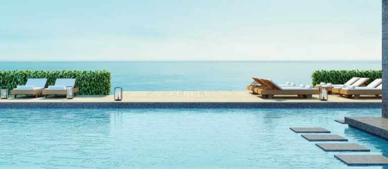 Infinity-Pool eines Luxushotels am Meer, umgeben von modernen Liegestühlen und grünen Hecken auf einer Holzterrasse. Im Vordergrund führt ein Weg aus Steinplatten durch das Wasser.
