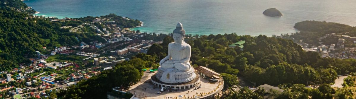 Grosse, weisse Buddha-Statue auf Hügel mit Blick auf bewaldete Landschaft und Küstenlinie. Der Ozean erstreckt sich im Hintergrund. Kleine Gebäude und Straßen säumen die Hänge im Vordergrund.