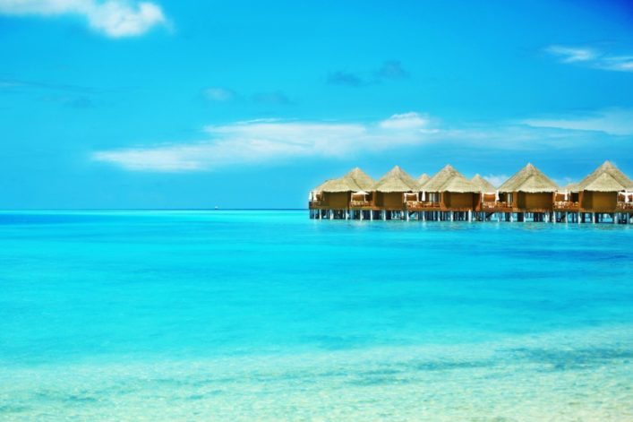 Türkisblaues Meer, klarer Himmel mit wenigen Wolken, auf der rechten Seite stehen strohgedeckte Wasserbungalows auf Stelzen, perfekte Kulisse für Flitterwochen auf den Malediven.