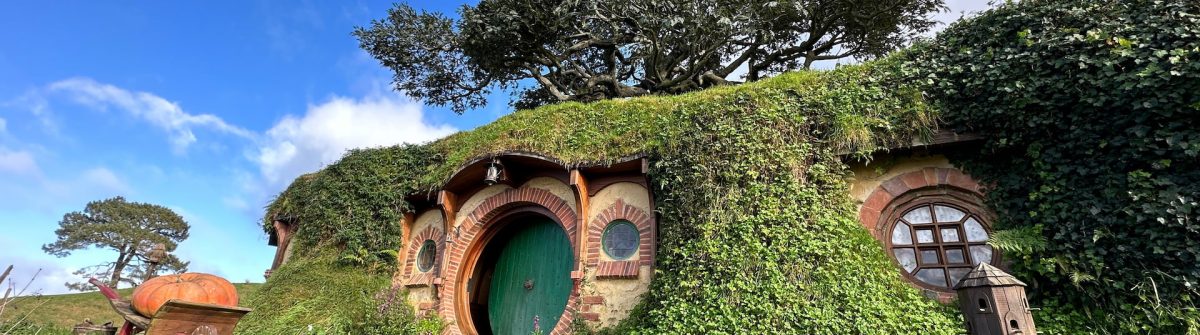 Eine typische Hobbit-Höhle in Hobbiton, Neuseeland. Rundherum sind saftig grüne Wiesen und im Hintergrund ein strahlend blauer Himmel mit vereinzelten Wolken.