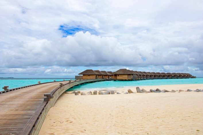 Ein Holzsteg führt über türkisblaues Wasser zu einer Reihe von strohgedeckten Strandvillen, umgeben von hellem Sand unter bewölktem Himmel – perfektes Ziel für Flitterwochen auf den Malediven.