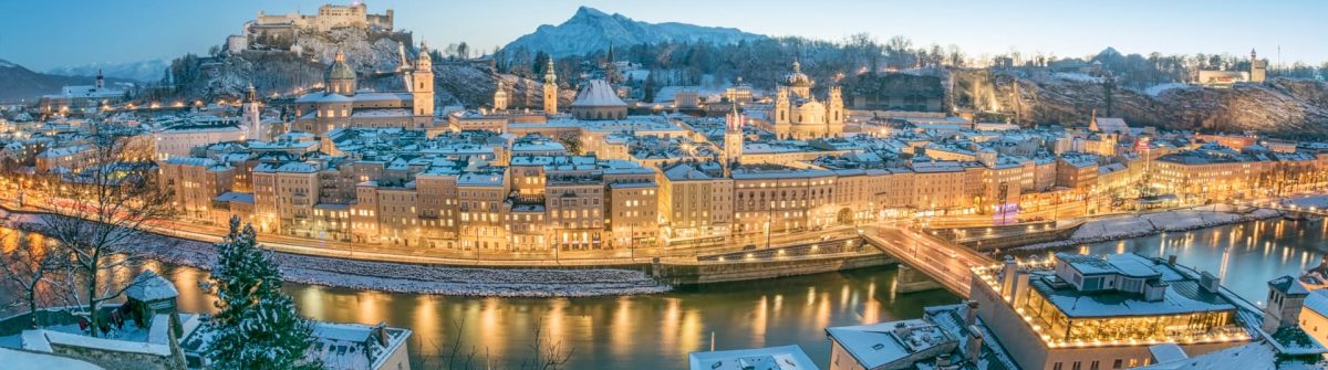 Winterliche Stadtansicht von Salzburg mit schneebedeckten Dächern, Hohensalzburg auf einem Hügel im Hintergrund, beleuchtete Gebäude entlang eines Flusses bei Abenddämmerung.