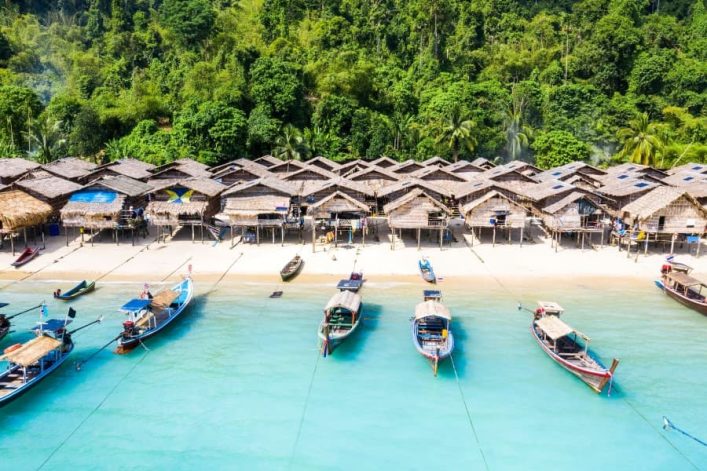 Strandhütten an einem smaragdgrünen Meer, umgeben von üppigem Grün, ähneln der Idylle der Surin-Inseln in Thailand, mit traditionellen Booten im Vordergrund.