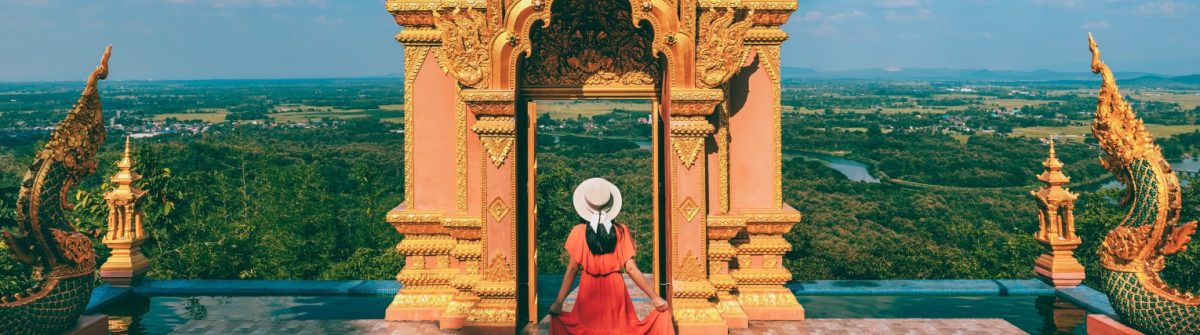 Eine Person in rotem Kleid und Hut steht vor einem reich verzierten goldenen Tor im Wat Phra That Doi Phra Chan Tempel, Lampang, mit Blick auf eine weite, bewaldete Landschaft.