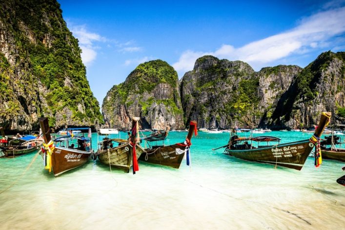Türkisblaues Meer mit traditionellen Langbooten am weißen Sandstrand von Koh Phi Phi, umgeben von majestätischen Kalksteinfelsen.