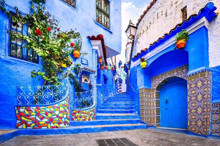 Eine malerische Gasse in Chefchaouen, geprägt von leuchtend blauen Häusern und Treppen. Bunte Blumentöpfe hängen an den Wänden, und links rankt sich ein grüner Strauch über ein Geländer.