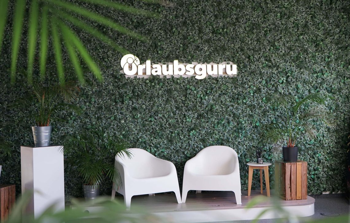 2 weiße Stühle vor einer Efeuwand mit dem Urlaubsguru-Logo