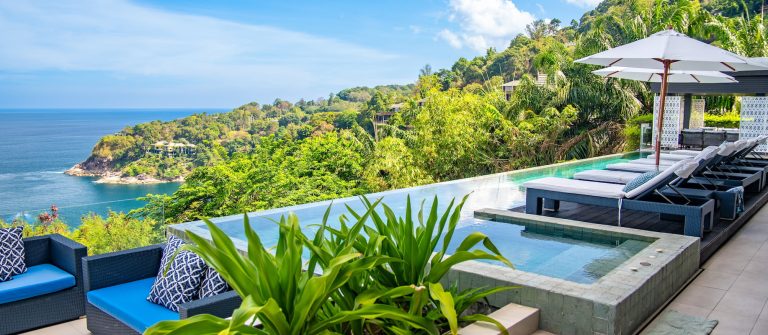 Ein Luxushotel auf Phuket. Blick über den Infinity-Pool aufs Meer. Der Himmel ist leicht bewölkt und ringsum sieht man saftig grüne Pflanzen.