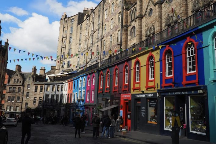 Bunte Häuserfassaden in kräftigen Farben säumen die kurvige Victoria Street in Edinburgh, einer beliebten Sehenswürdigkeit. Oben hängen Wimpelketten, Menschen flanieren auf dem Kopfsteinpflaster.