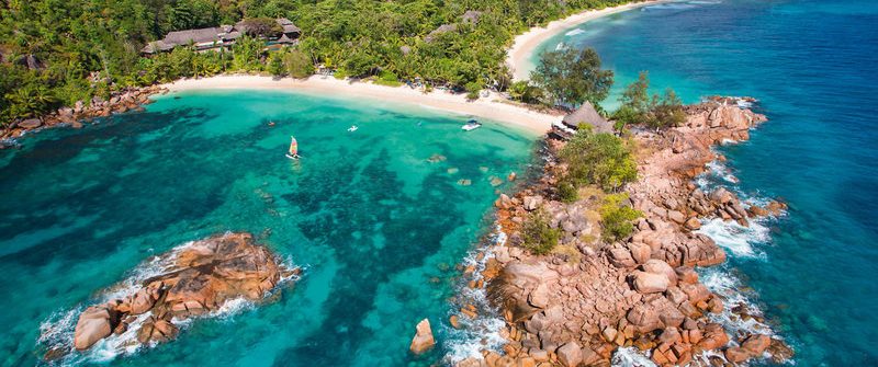Türkises Wasser umgibt felsige Inselgruppe mit üppiger Vegetation. Ein Segelboot links im Wasser. Im Hintergrund dichter Wald und geschwungener Sandstrand unter blauem Himmel.