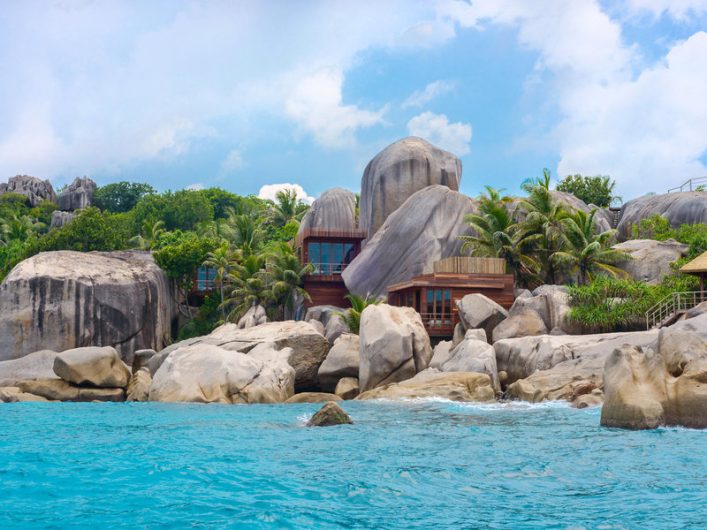 Holzhäuser eingebettet in üppige grüne Palmen und große Granitfelsen, die zum türkisblauen Meer hin abfallen, unter einem blauen Himmel mit leichten Wolken.