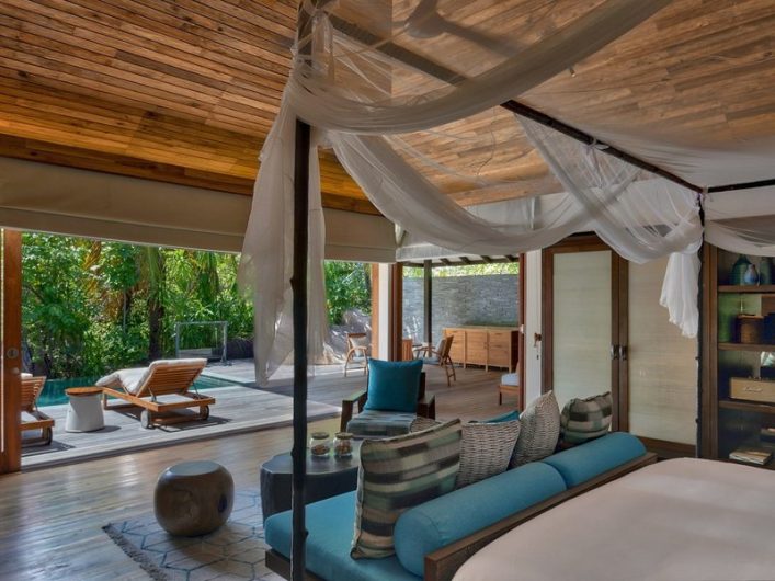 Luxuriöser, offener Raum mit Holzdecke, Himmelbett und blauen Kissen. Große Glastüren führen zu einer Terrasse mit Pool, Liegestühlen und üppigem Grün im Hintergrund. Regal mit Deko rechts.